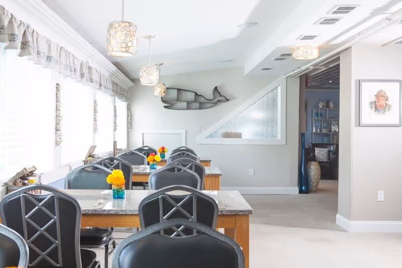 Bright dining area with multiple tables and black chairs arranged in rows. Each table has a small vase with colorful flowers. Large windows with sheer curtains allow natural light to fill the room. Decorative pendant lights hang from the ceiling. A wall features a metal whale sculpture and a framed portrait is visible on the adjacent wall. The space has a clean, welcoming atmosphere.