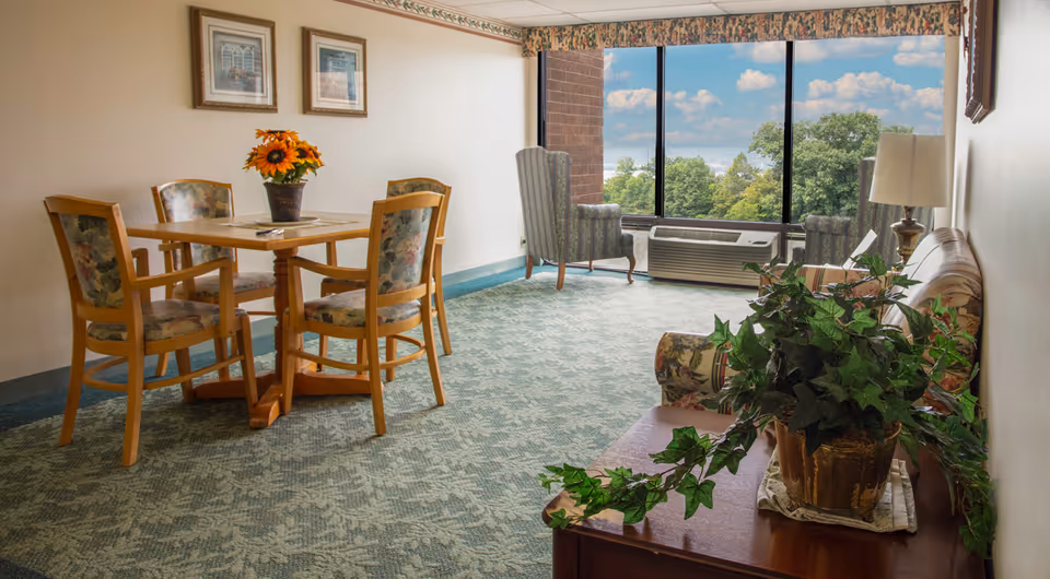 A cozy senior living common area with a wooden table and four floral upholstered chairs on the left, two striped armchairs near a large window showing a view of trees and blue sky, a floral patterned sofa on the right, a wooden side table with a potted plant, and framed artwork on the wall.