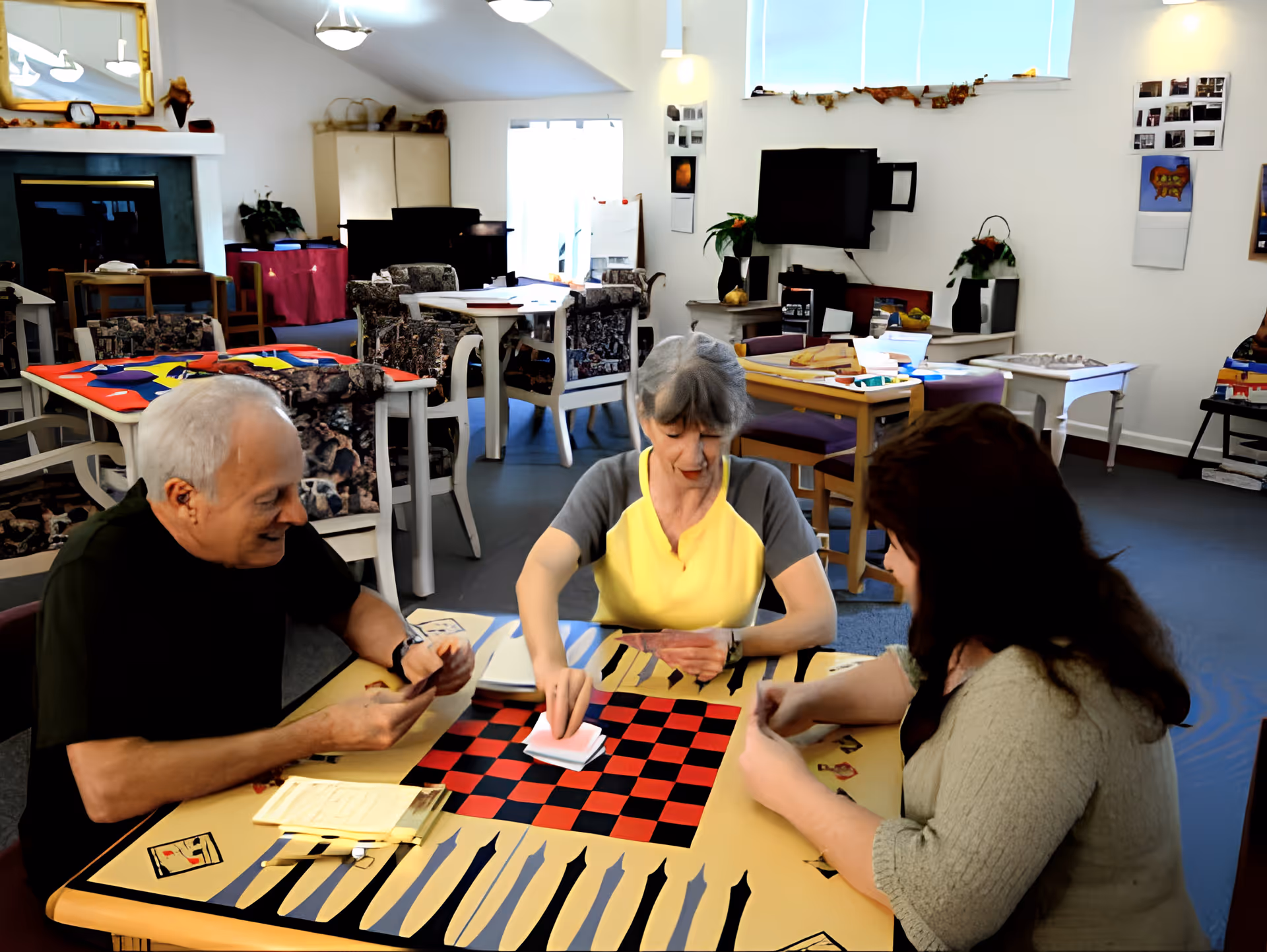 Three elderly people sitting around a table playing a card game in a well-lit common room with multiple tables and chairs, a fireplace, and a television mounted on the wall.