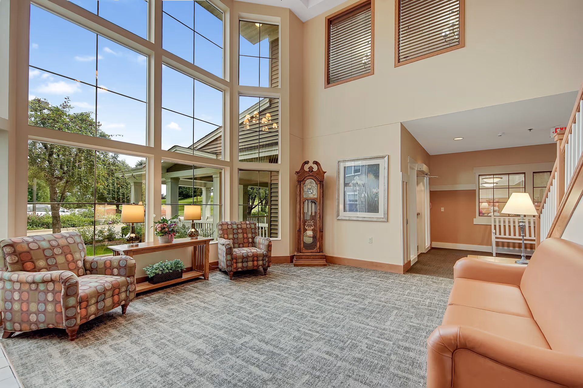 A spacious and well-lit living room area with large floor-to-ceiling windows showing an outdoor view with trees and a building exterior. The room features two patterned armchairs, a wooden console table with lamps and plants, a grandfather clock, a framed picture on the wall, a leather sofa, and a staircase leading to an upper floor.