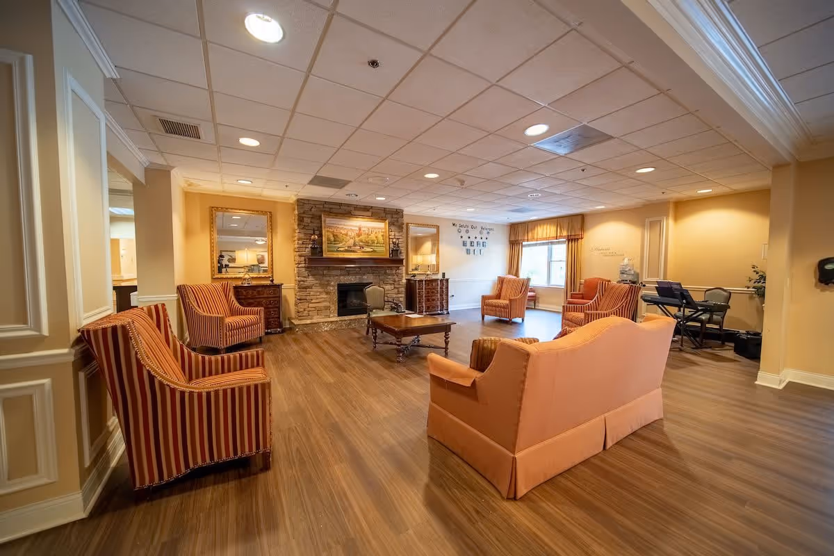 A spacious senior living facility common area with wooden flooring, several upholstered armchairs and a sofa arranged around a wooden coffee table. The room features a stone fireplace with a painting above it, two large mirrors on either side, and a window with curtains letting in natural light. There is also a keyboard and chair in the corner, and soft yellow walls with recessed lighting in the ceiling.