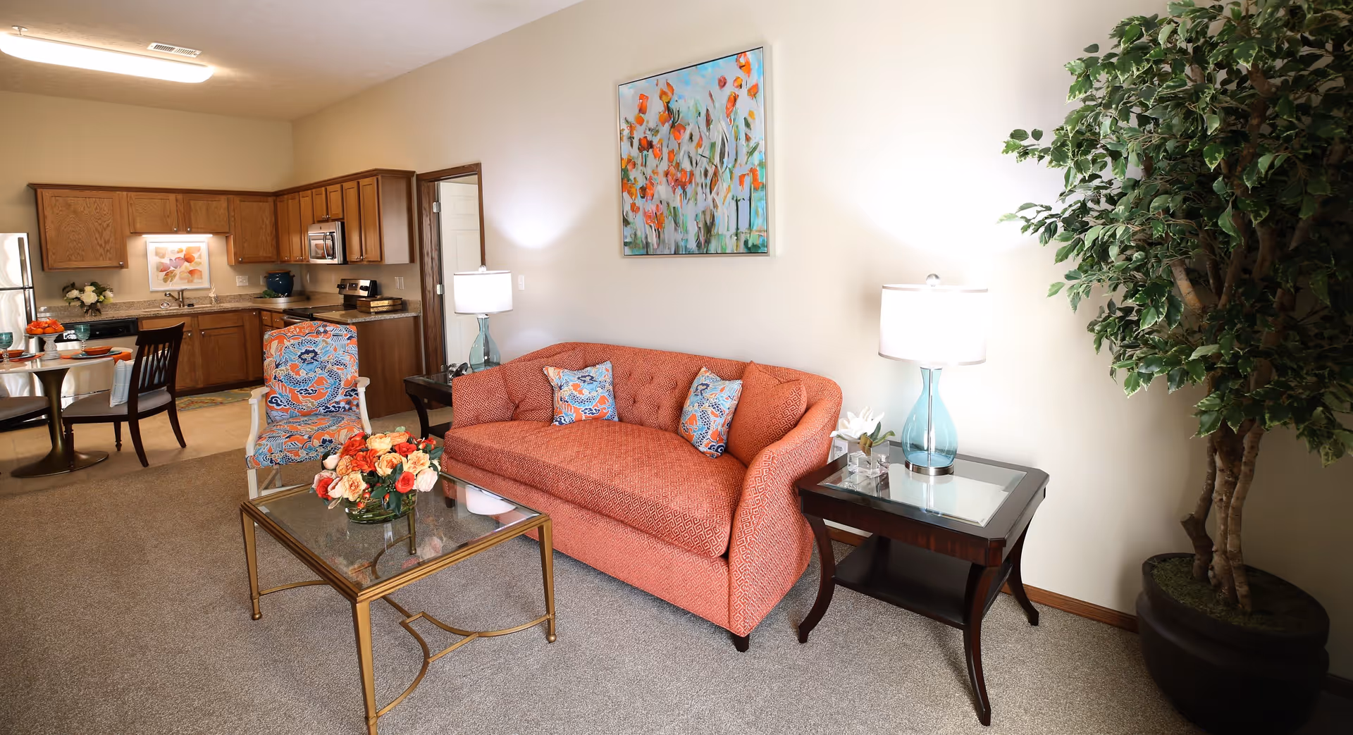 A cozy living room area with a coral-colored sofa adorned with patterned pillows, a glass coffee table with a floral arrangement, a patterned armchair, two side tables with lamps, a large potted plant, and a colorful painting on the wall. In the background, there is an open kitchen with wooden cabinets and a dining table with chairs.