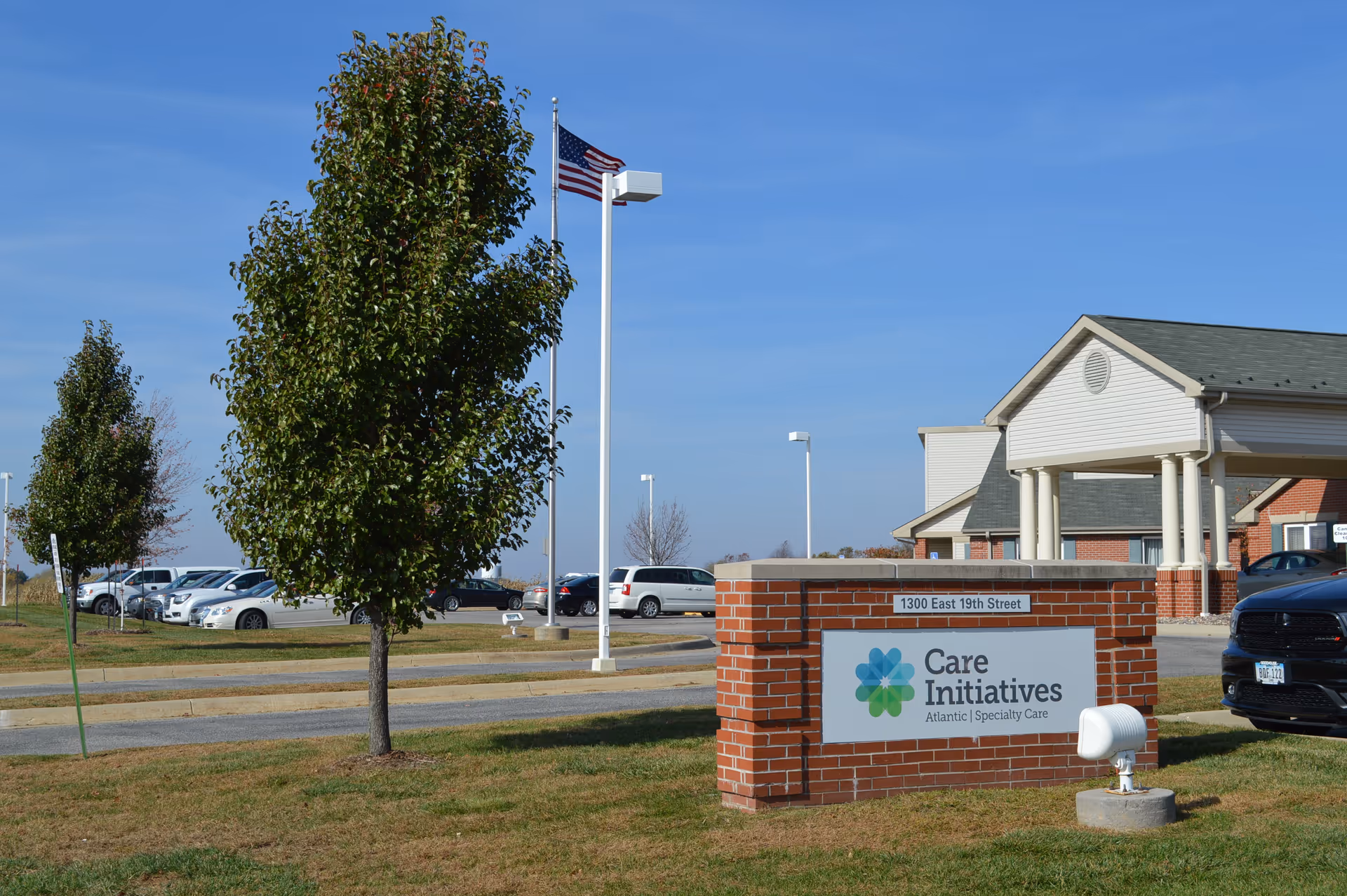Exterior view of a senior care facility named Care Initiatives Atlantic Specialty Care, showing a brick sign with the facility name and address 1300 East 19th Street, a parking lot with cars, a building entrance with columns, trees, and an American flag on a flagpole against a clear blue sky.