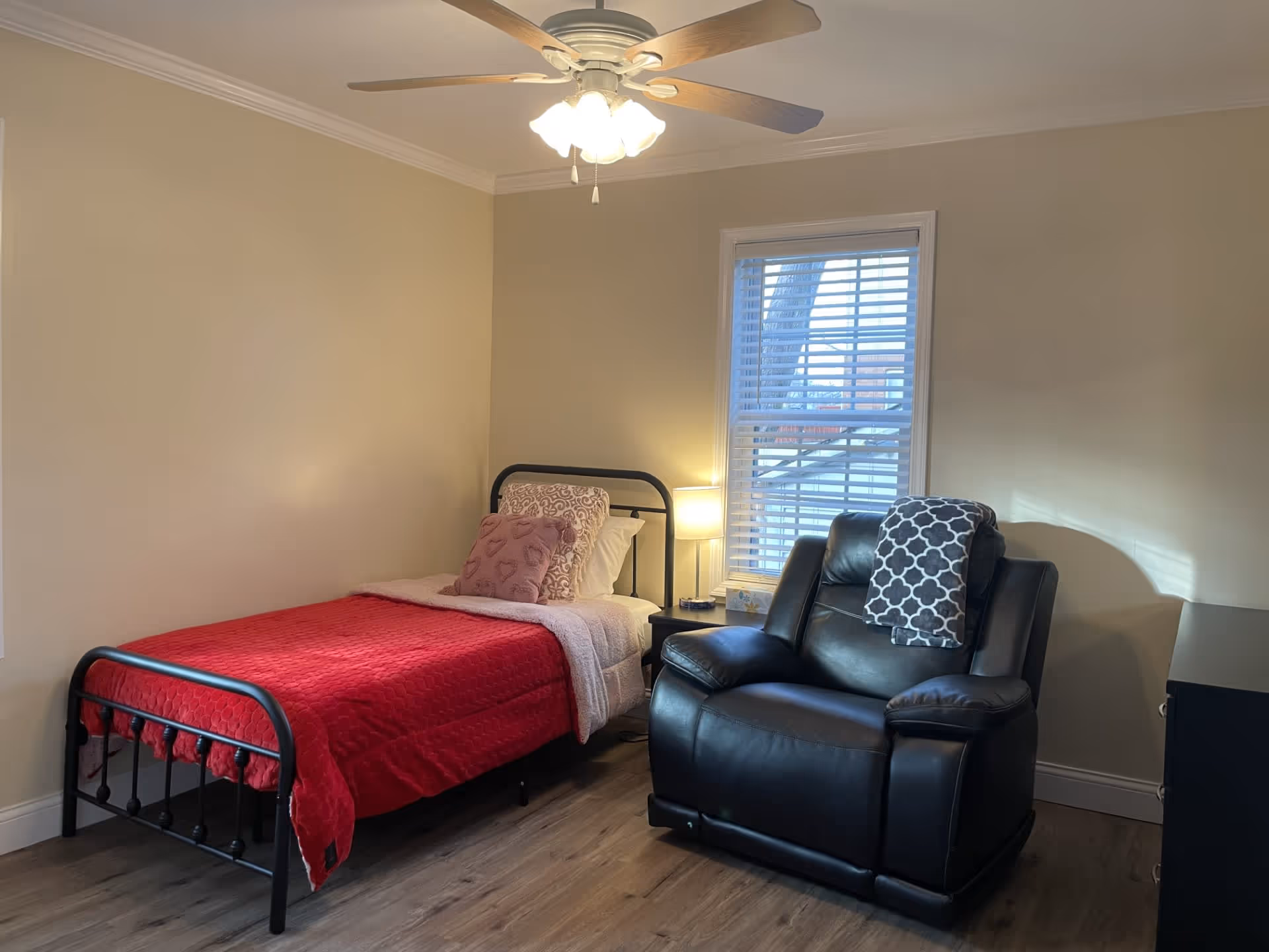 A cozy bedroom with a single black metal bed frame covered with a red blanket and decorative pillows. Next to the bed is a small nightstand with a lamp and a box of tissues. A large black leather recliner chair with a patterned throw blanket is positioned near a window with white blinds. The room has beige walls, wood flooring, and a ceiling fan with lights.