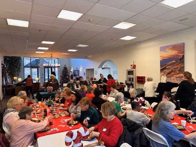 A large group of elderly people gathered in a spacious dining room decorated for the holidays, sitting at tables covered with red and green tablecloths, eating and socializing. There is a Christmas tree in the background and festive decorations on the tables.