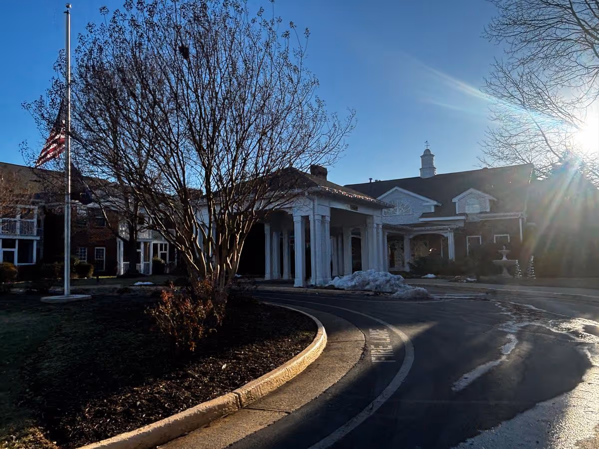 Front entrance of a senior living facility with a covered portico, circular driveway, flag at half-mast, and bare trees in bright sunlight.