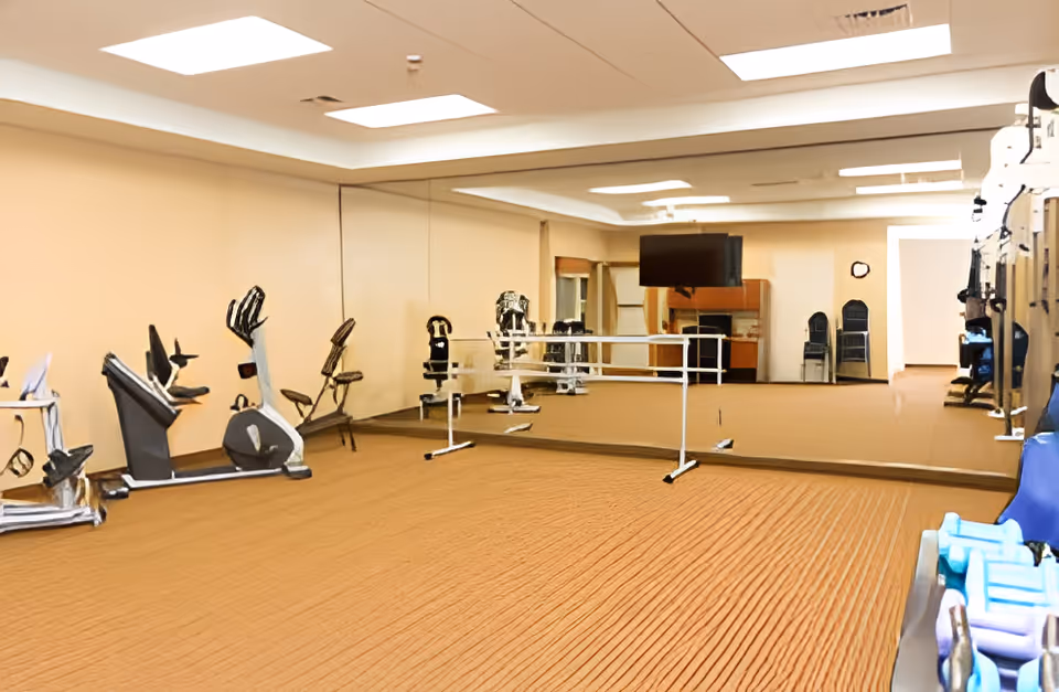 A fitness room with exercise equipment including stationary bikes and weight machines. The room has a large wall mirror, a ballet barre, a television mounted on the wall, and a carpeted floor. There are also some stacked chairs and a clock on the far wall.