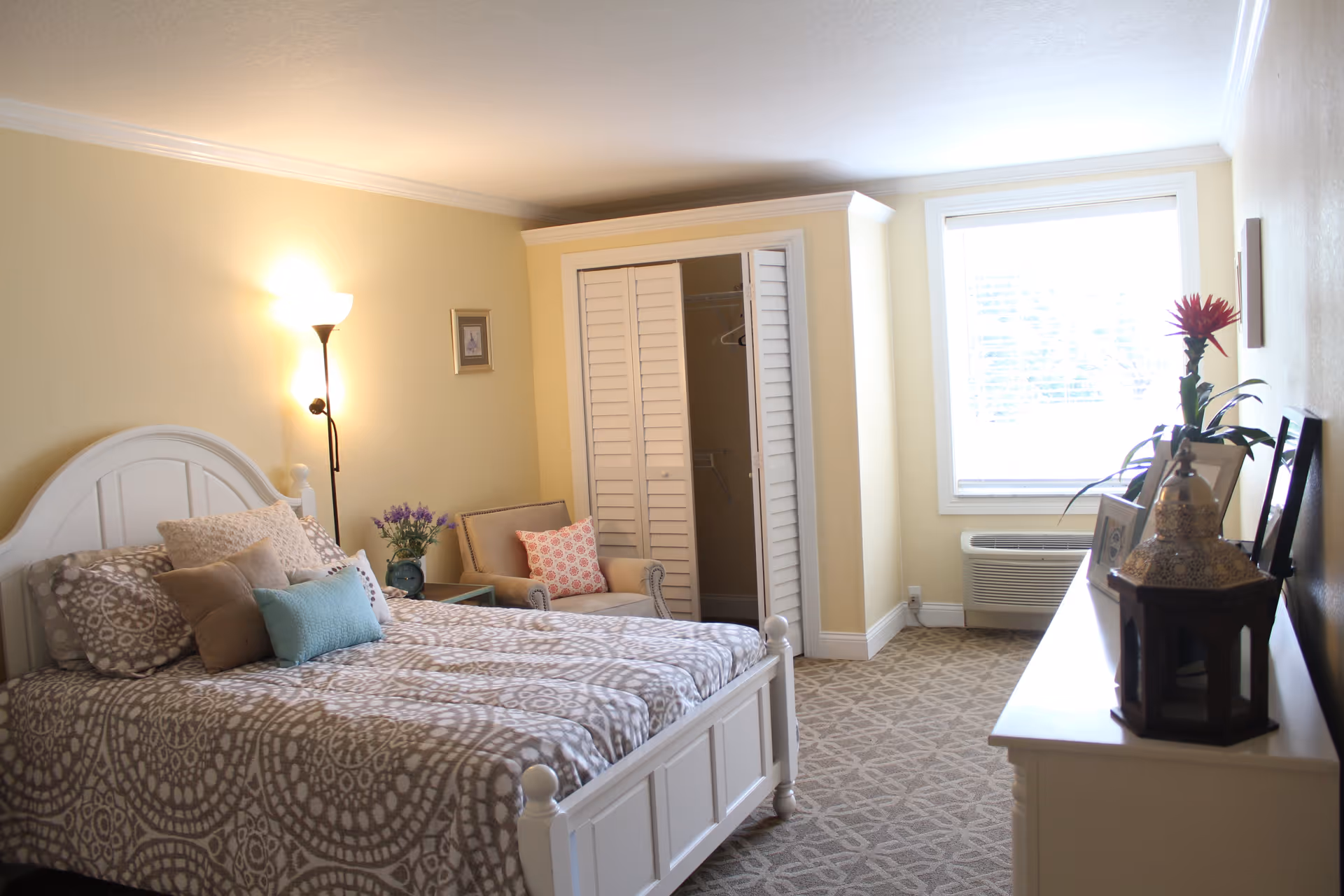 A cozy bedroom in a senior living facility with a white wooden bed frame, patterned bedding, several pillows, a floor lamp, an armchair with a decorative pillow, a small side table with a vase of flowers, an open closet with white shutter doors, a window with blinds, and a white dresser topped with framed pictures and a decorative lantern.