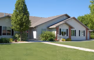Single-story residential building with light-colored siding and brick accents, surrounded by a well-maintained lawn and trees under a clear blue sky.