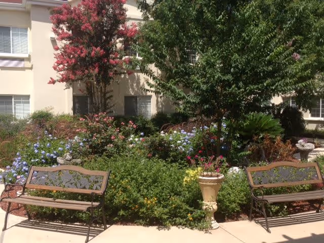 Outdoor garden area with two metal benches facing each other, surrounded by lush greenery, colorful flowers, and trees. The background shows a beige building with windows.