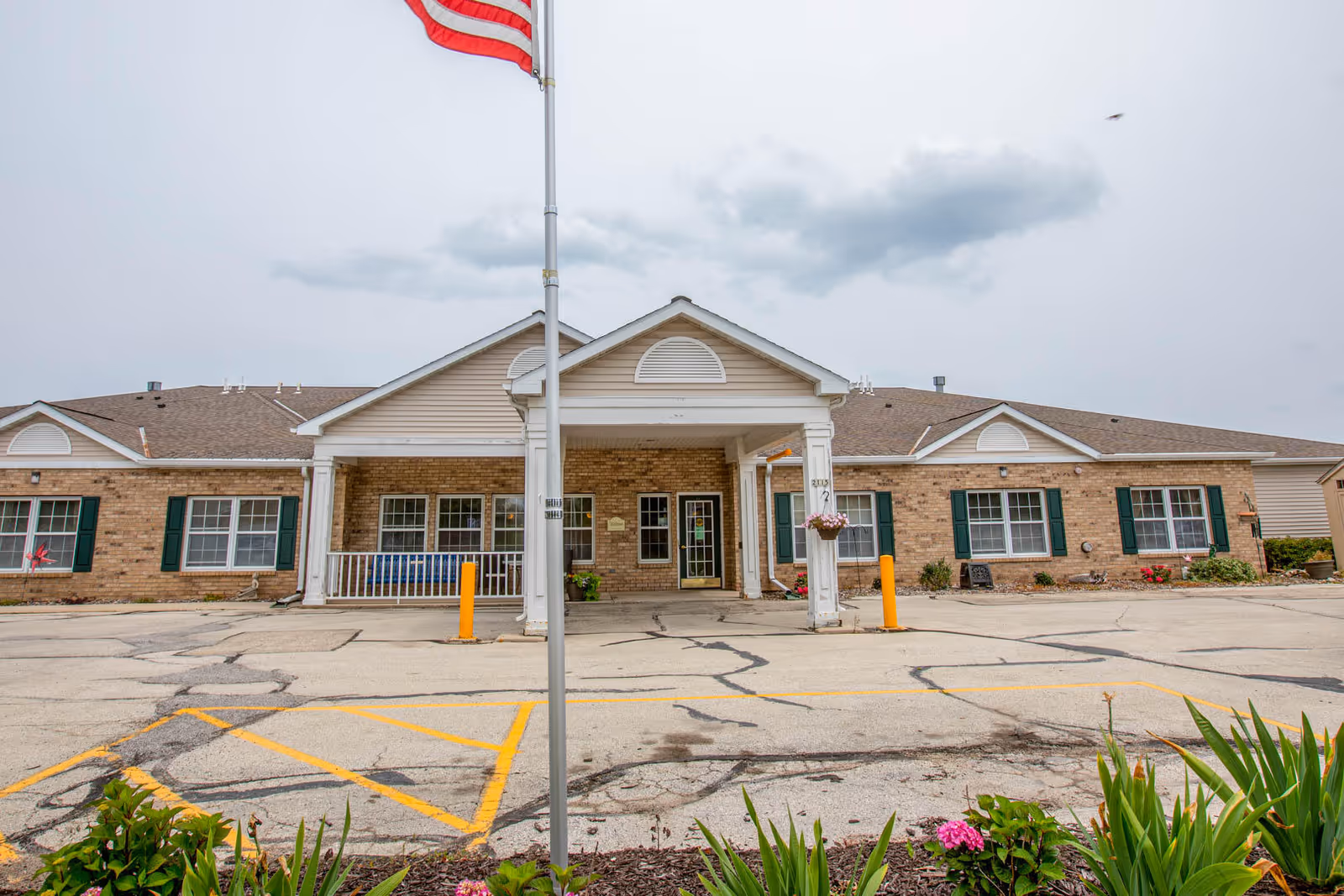 Front entrance of a single-story brick assisted living building with a covered portico, an American flag on a pole, parking area, and landscaping.