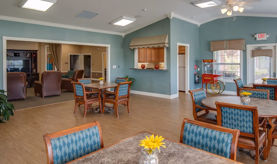 A bright and spacious common area in an assisted living facility featuring multiple round tables with blue patterned chairs and small yellow flower centerpieces. The room has light wood flooring, teal walls, and large windows letting in natural light. In the background, there is a cozy seating area with brown leather armchairs and a TV. A popcorn machine is visible near the windows, and an exit door is on the right side.