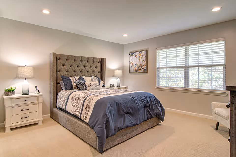 A neatly made bed with a tufted gray headboard in a bedroom. The bed has patterned bedding in shades of blue and white with multiple pillows. There are two white nightstands on either side of the bed, each with a lamp. A window with white blinds is on the right wall, and a framed floral painting hangs above the right nightstand. The room has beige carpeting and recessed ceiling lights.