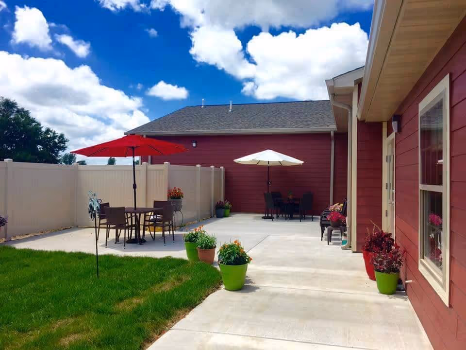 Outdoor patio area with two seating arrangements, each with a table and umbrella, surrounded by potted plants and a white privacy fence. The building exterior is red with beige trim, and the sky is partly cloudy with blue patches.