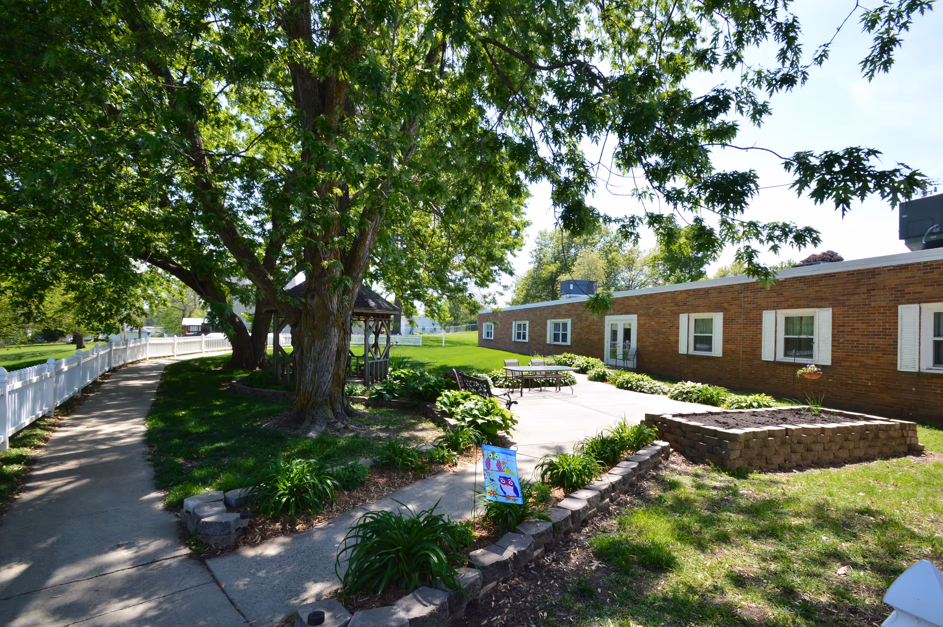 Outdoor patio area at Bedford Specialty Care featuring a paved walkway, a white fence, large leafy trees providing shade, a gazebo, garden beds, and a brick building with white-framed windows in the background.