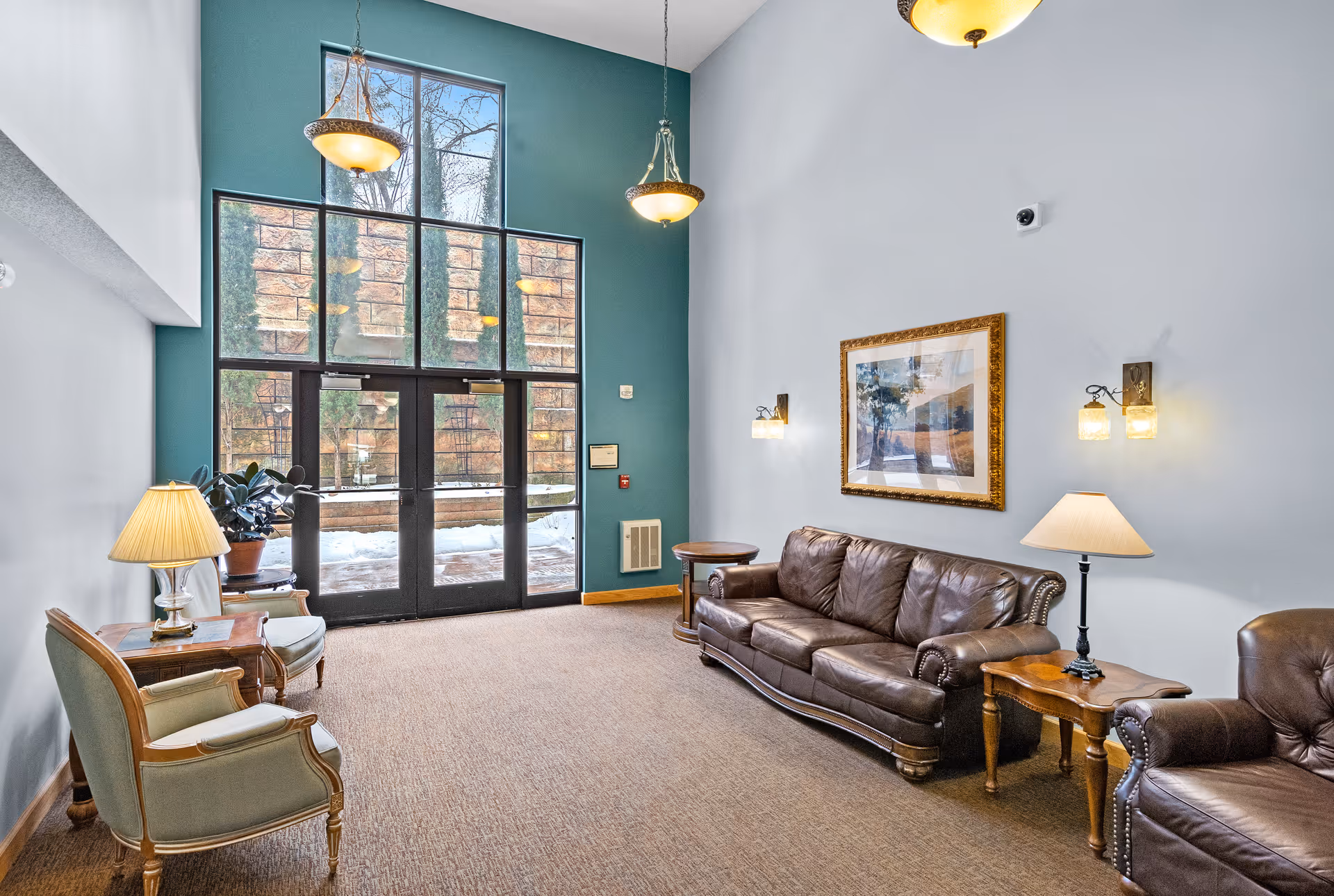 A senior living facility common area with high ceilings and large windows letting in natural light. The room features a brown leather sofa, matching armchair, two wooden side tables with lamps, two upholstered chairs with a small wooden table and lamp, and a framed landscape painting on the wall. Outside the glass doors, snow is visible.