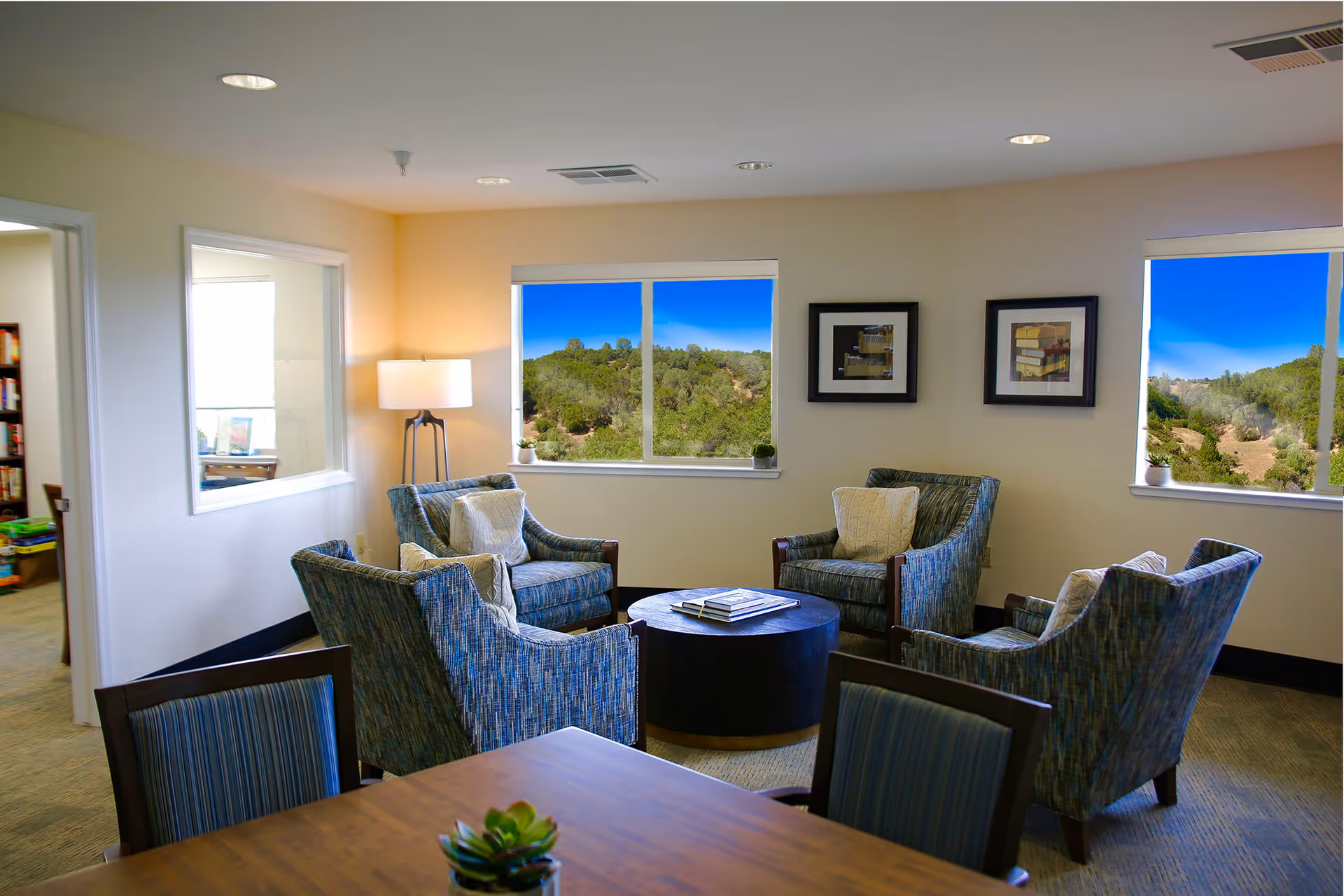 A cozy sitting area in a senior living facility with four blue patterned armchairs arranged around a round black coffee table. The room has beige walls, two windows showing a scenic view of green hills and blue sky, a floor lamp in the corner, and two framed pictures on the wall. A wooden table with a small plant is partially visible in the foreground.