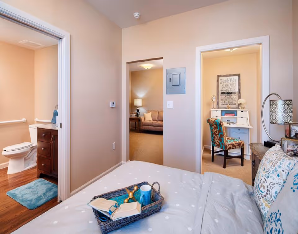 View from a bedroom showing a bed with a tray holding a book, glasses, and a cup. To the left is an open door leading to a bathroom with a toilet, vanity, and blue bath mat. Straight ahead is a doorway to a living room with a sofa and lamp. To the right is a small office area with a white desk, a colorful chair, and a framed quote on the wall.