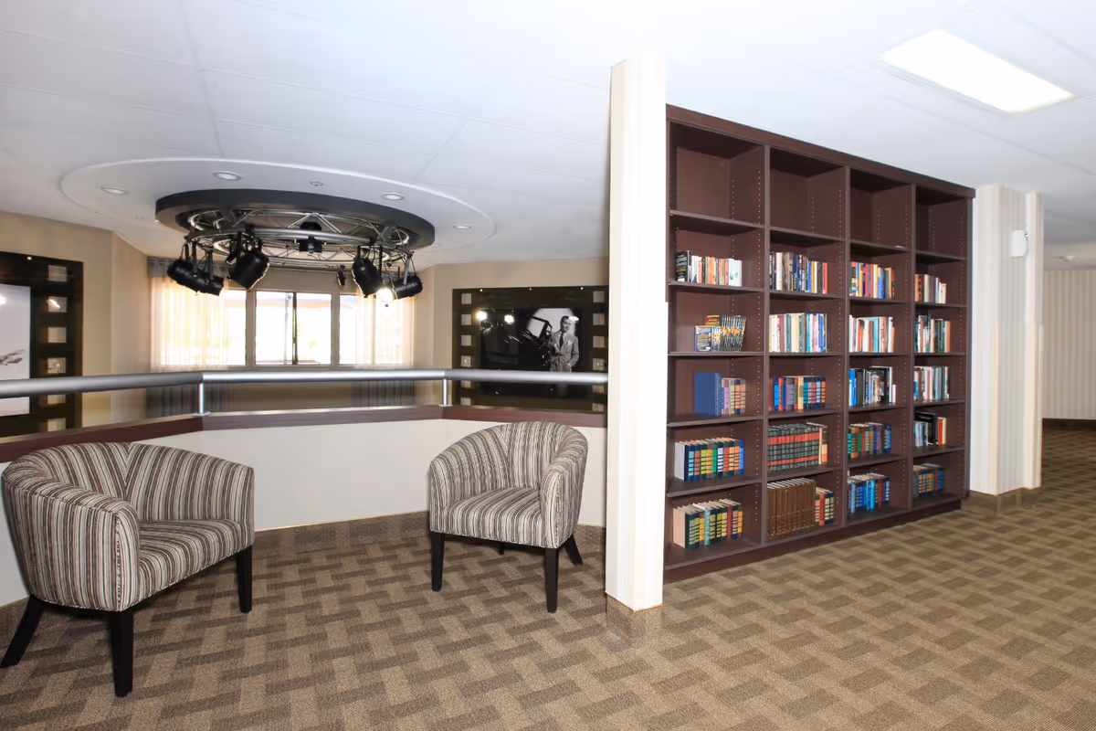 Interior view of a senior living facility lounge area with two striped armchairs and a large dark wood bookshelf filled with books. The space has patterned carpet flooring, a railing overlooking a lower level, and ceiling lights with mounted spotlights.