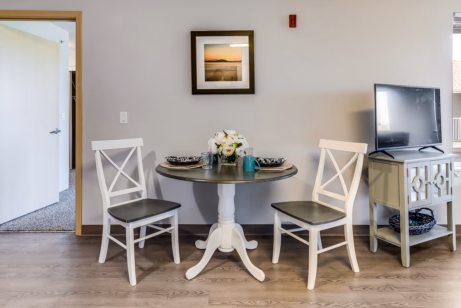 Small dining area with a round table set for two, two white chairs, a TV on a side table, and framed artwork on the wall.