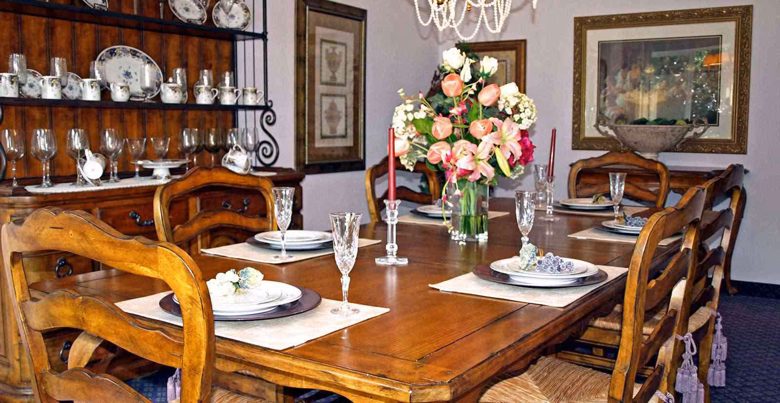 A wooden dining table set for six with plates, crystal glasses, and red candles in glass holders. A vase with a colorful bouquet of flowers is placed in the center of the table. Behind the table, there is a wooden cabinet displaying fine china and glassware. The room has framed artwork on the walls and a chandelier hanging above the table.