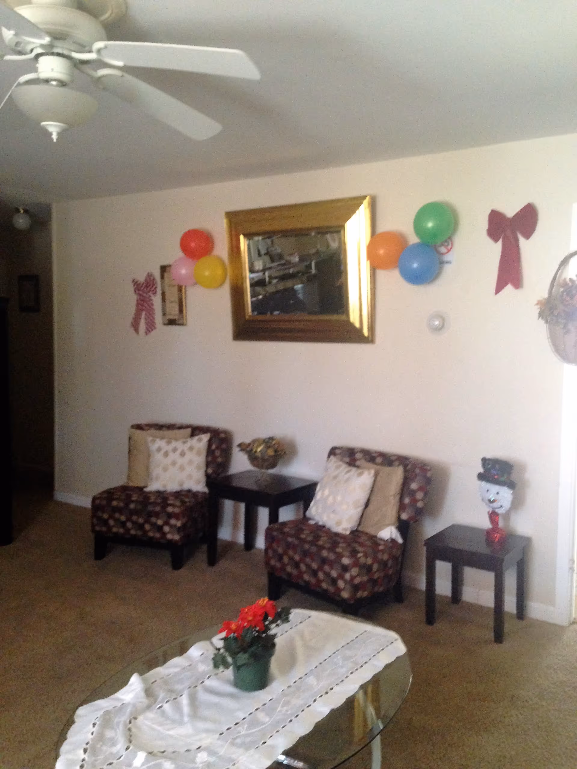Cozy living room with two patterned chairs and side tables, a glass coffee table with a potted plant, a ceiling fan, a wall mirror and festive balloons on the wall.