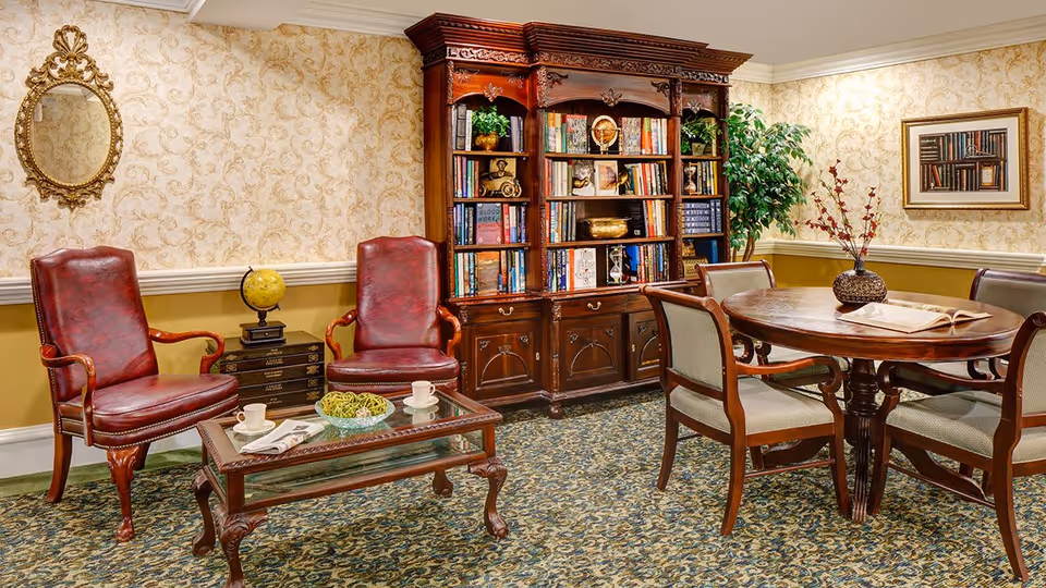 A cozy sitting area in a senior living facility featuring two red leather armchairs with wooden frames, a glass coffee table with two cups and a decorative bowl, a wooden bookshelf filled with books and decorative items, a round wooden table with four chairs, a potted plant, and framed artwork on the wall. The room has patterned wallpaper and carpeted flooring.