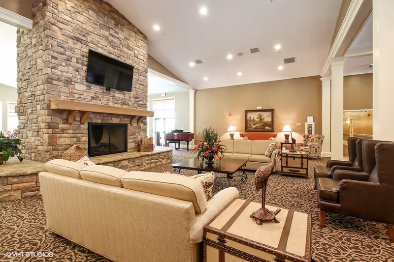 A spacious senior living facility common area with beige sofas, patterned carpet, a stone fireplace with a mounted flat-screen TV, a coffee table with a floral arrangement, and additional seating including leather chairs and an armchair. The room is well-lit with recessed ceiling lights and has a warm, inviting atmosphere.