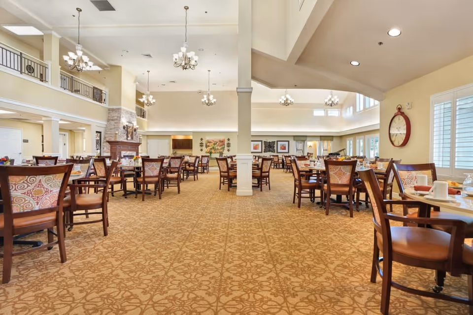 Spacious senior dining room with multiple wooden tables and upholstered chairs, chandeliers, a fireplace and large windows.
