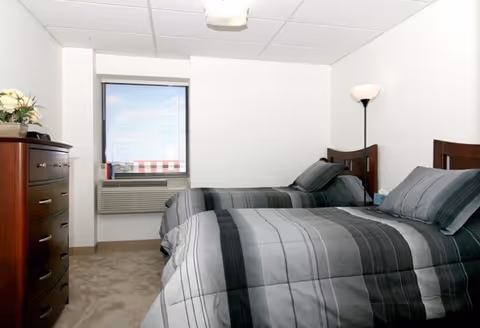 A simple bedroom with two twin beds featuring gray and black striped bedding. There is a wooden dresser with a flower arrangement on top to the left, a window with blinds and an air conditioning unit below it, and a floor lamp in the corner to the right.