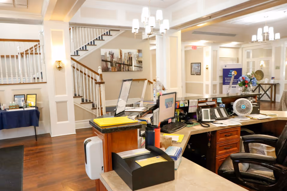 Reception desk and lobby area with a staircase, workstations, phones, and seating in a senior living facility.