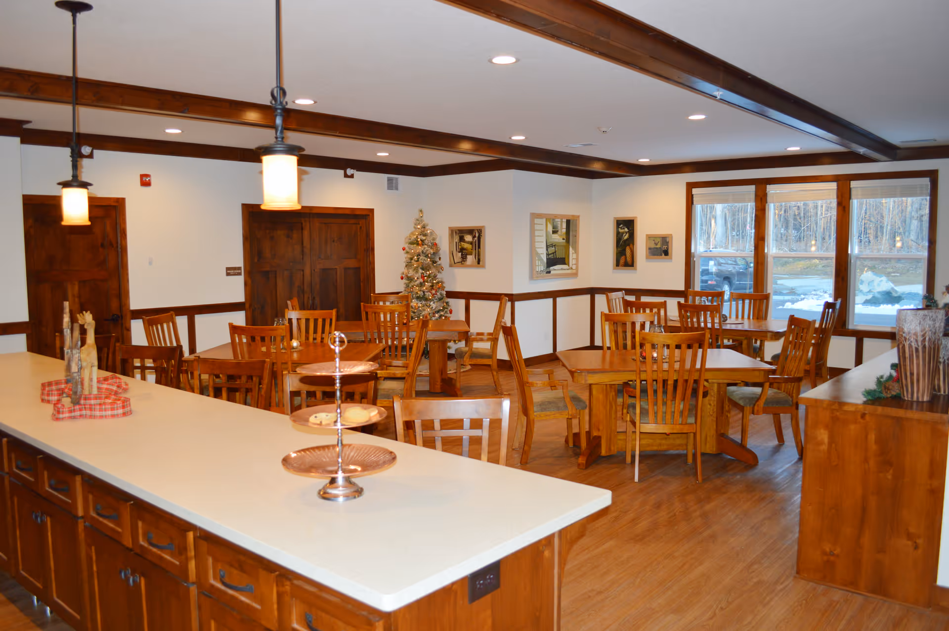 A cozy dining room with several wooden tables and chairs arranged neatly. There is a decorated Christmas tree in the corner, large windows showing a snowy outdoor scene, and warm lighting from ceiling fixtures. The room features wooden beams on the ceiling and wood-paneled lower walls.