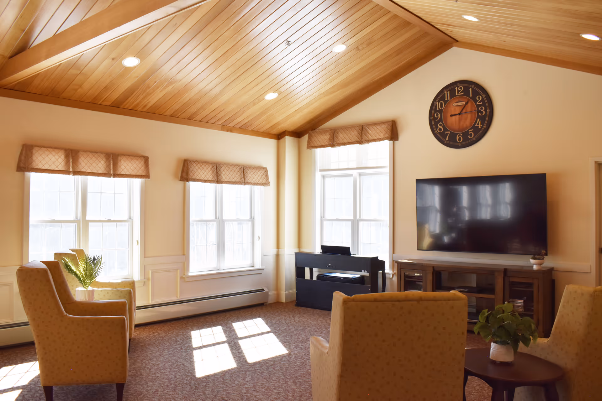 Sunlit common living room with upholstered chairs, large windows, a wall-mounted TV, and a wooden vaulted ceiling.