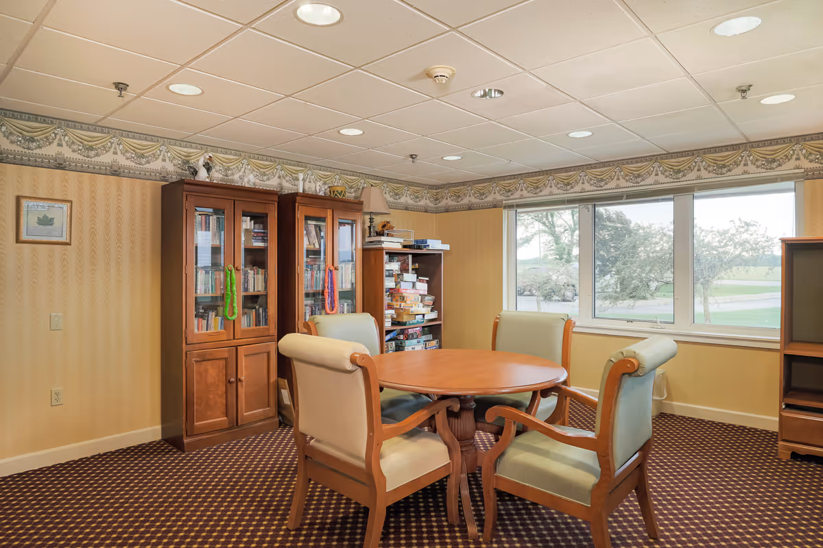 A cozy room with a round wooden table surrounded by four cushioned chairs. Behind the table, there are two wooden cabinets filled with books and board games. A large window lets in natural light and offers a view of trees and greenery outside. The room has patterned carpet and wallpaper with a decorative border near the ceiling.