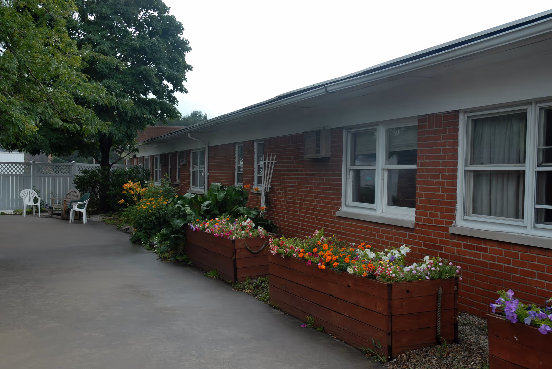 Brick single-story care facility exterior with windows, wooden planter boxes of colorful flowers, a paved courtyard and chairs under a tree.