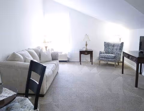 Bright carpeted living room with a beige sofa, patterned armchair, side tables with lamps, and a TV on a console.