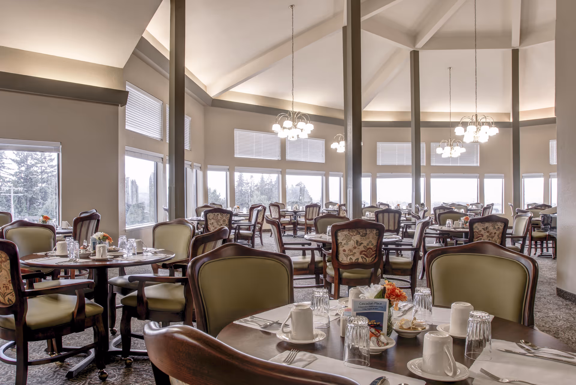 Spacious dining room with round tables and upholstered chairs set for a meal beneath chandeliers and large windows.