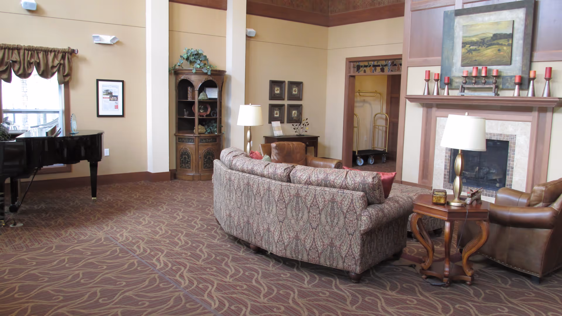 A cozy living room area with a patterned sofa, two leather armchairs, a wooden side table with a lamp, a fireplace decorated with candles and a painting above it, a wooden cabinet with plants, and a grand piano near a window with brown curtains.