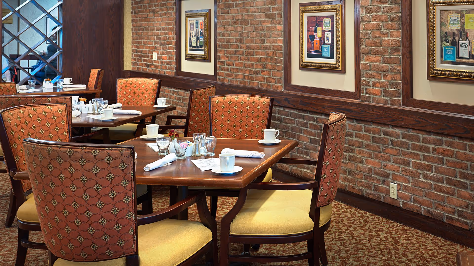 Dining area with wooden tables and upholstered chairs featuring a patterned red fabric and yellow seats. The room has exposed brick walls adorned with framed artwork depicting bottles. Tables are set with white cups, glasses, napkins, and condiments.