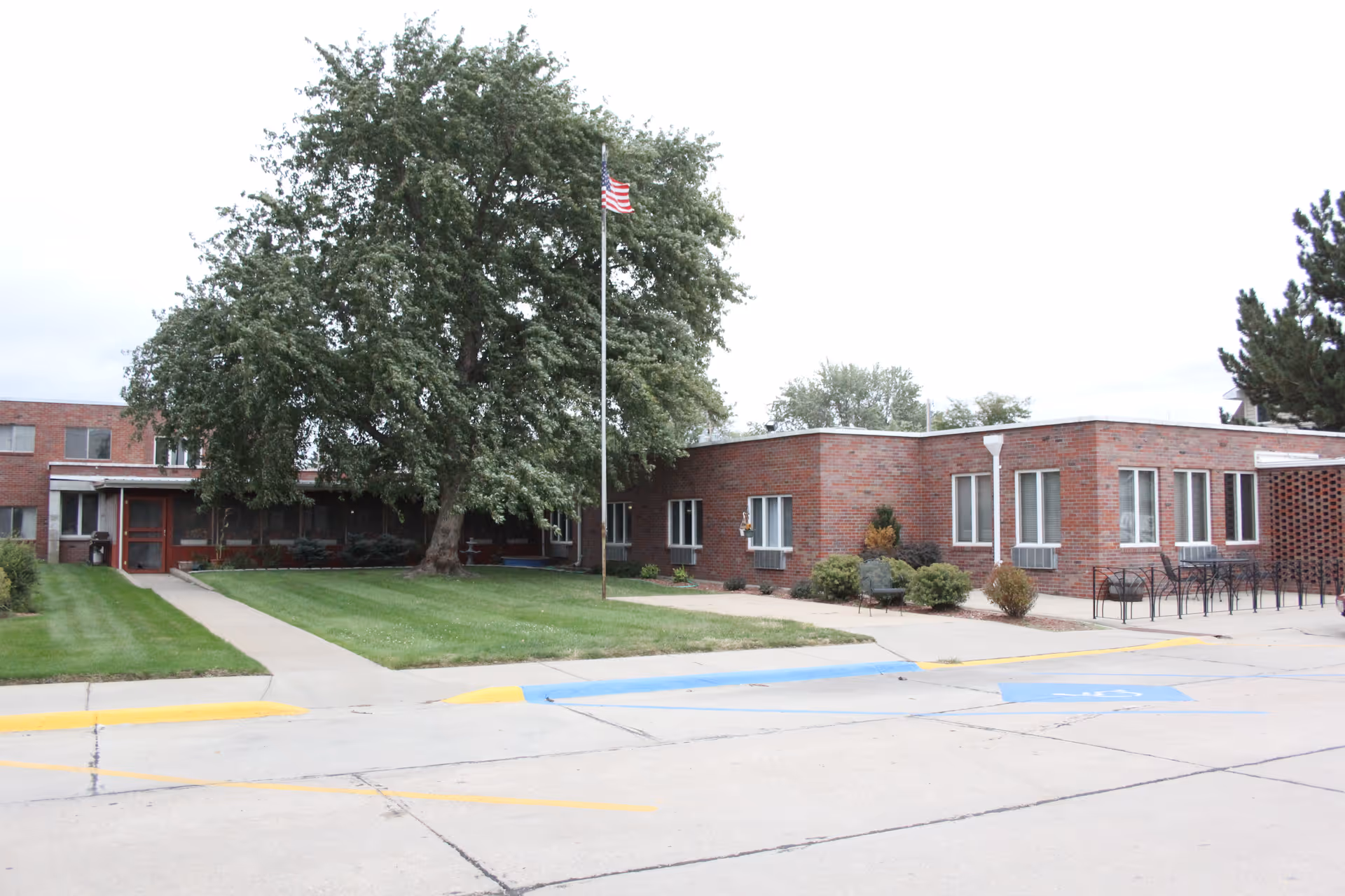 Front view of a single-story brick senior living building with a large tree, flagpole, lawn, and walkway to the entrance.