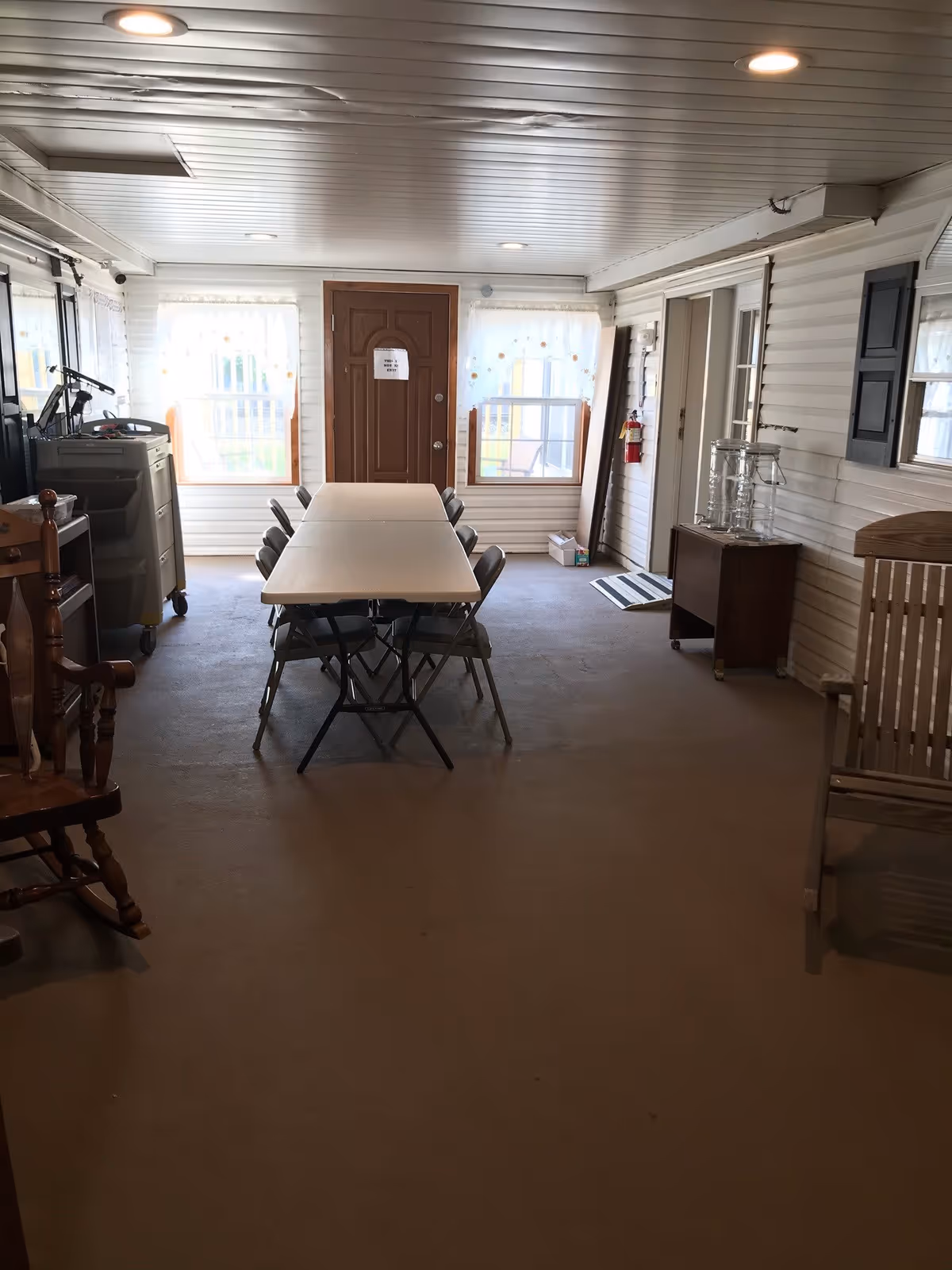 Interior room with a long folding table and several folding chairs arranged around it. The room has white paneled walls, two windows with sheer curtains, a wooden door with a sign, a wooden rocking chair, a small cabinet with glass containers, and a fire extinguisher mounted on the wall.