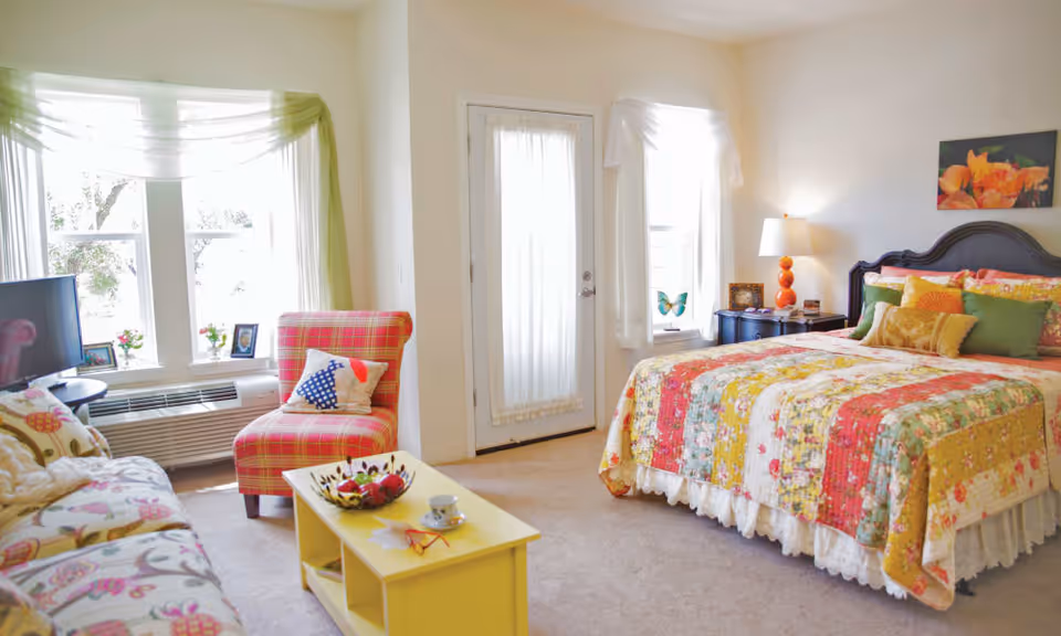 A bright and cozy bedroom with a colorful patchwork quilt on the bed, a bedside table with a lamp, a red plaid armchair with a decorative pillow, a floral patterned sofa, a yellow coffee table with a cup and decorative items, and large windows with sheer curtains letting in natural light.