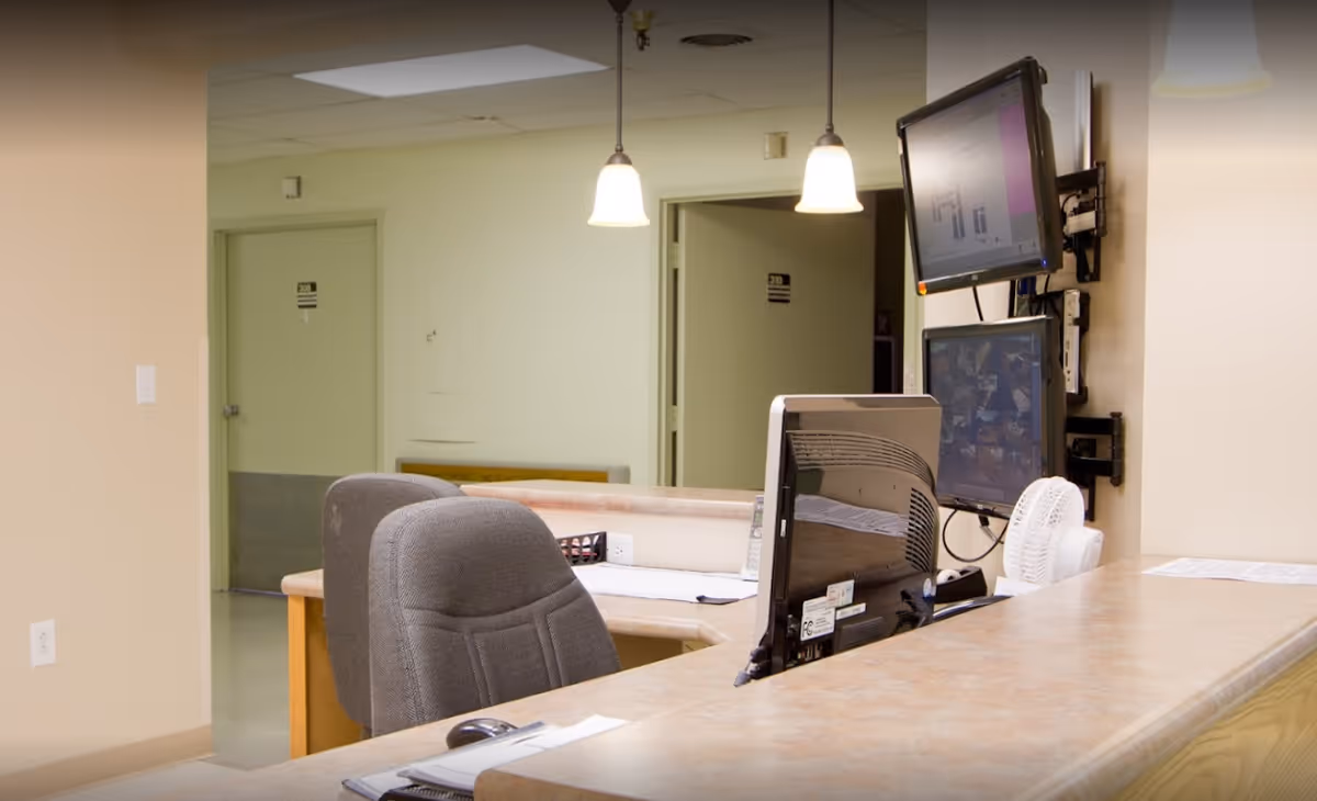 Reception or nurse station area with two gray office chairs, a countertop with computer monitors, a small fan, and two hanging pendant lights. Doors and hallway are visible in the background.