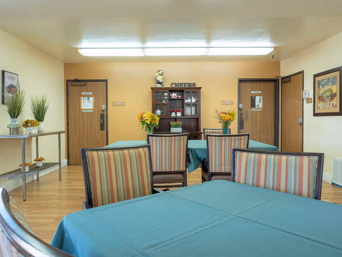 A dining room with tables covered in teal tablecloths and striped chairs. The room has wooden flooring and beige walls. There is a wooden cabinet against the far wall with decorative items and the word 'CHEERS' displayed on top. Two doors are visible on the back wall, along with framed artwork and vases with flowers and plants on a metal shelf to the left.