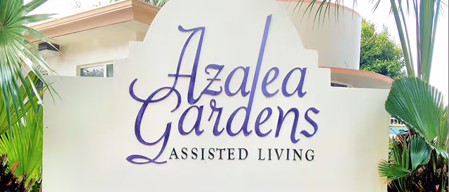 Outdoor sign reading 'Azalea Gardens Assisted Living' mounted on a light-colored wall with palm plants around it.