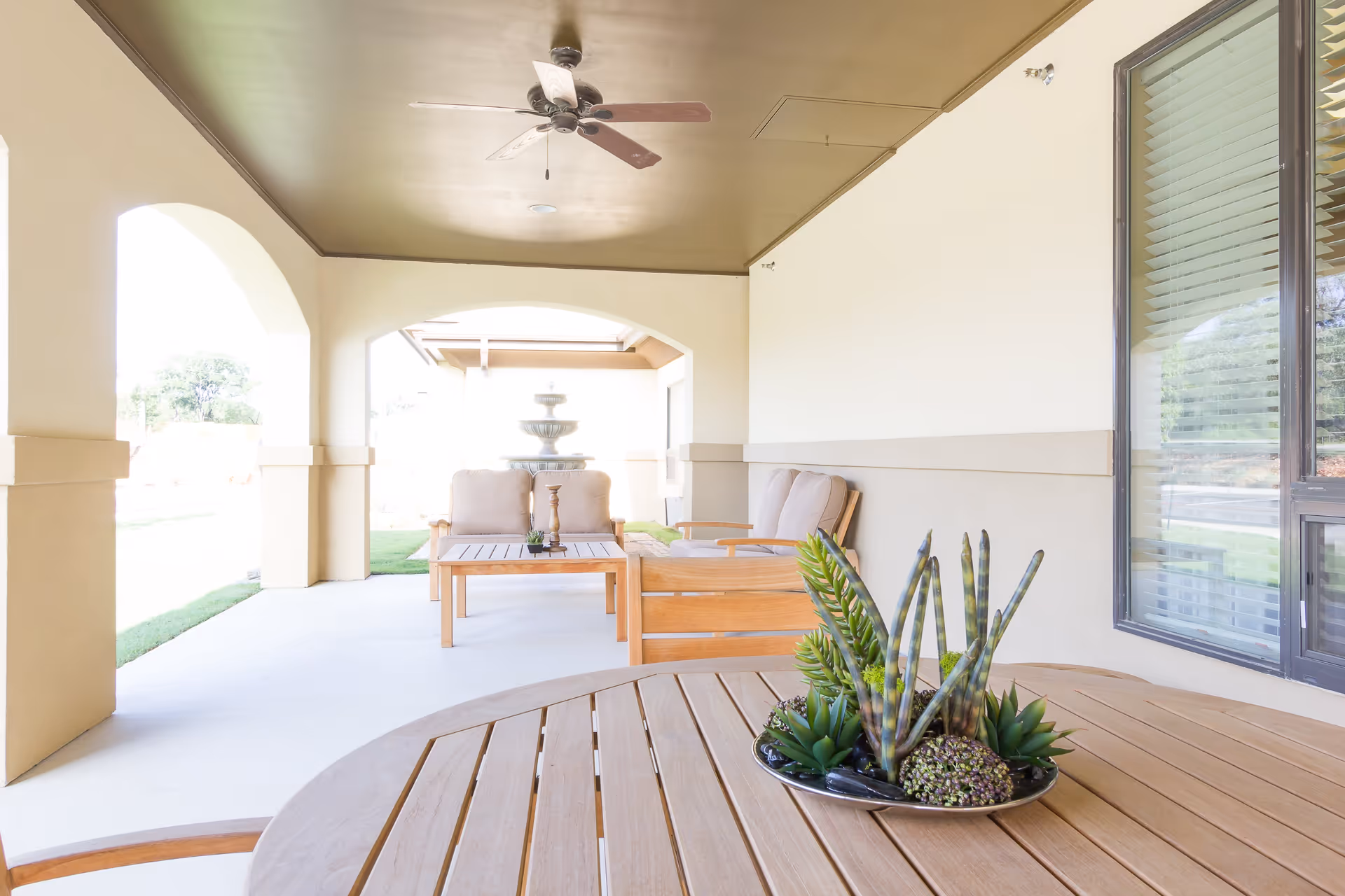 Covered outdoor patio area with wooden tables and cushioned chairs. A ceiling fan is mounted on the ceiling, and a decorative plant centerpiece is on the round table in the foreground. In the background, there is a water fountain and green lawn visible through the arches.