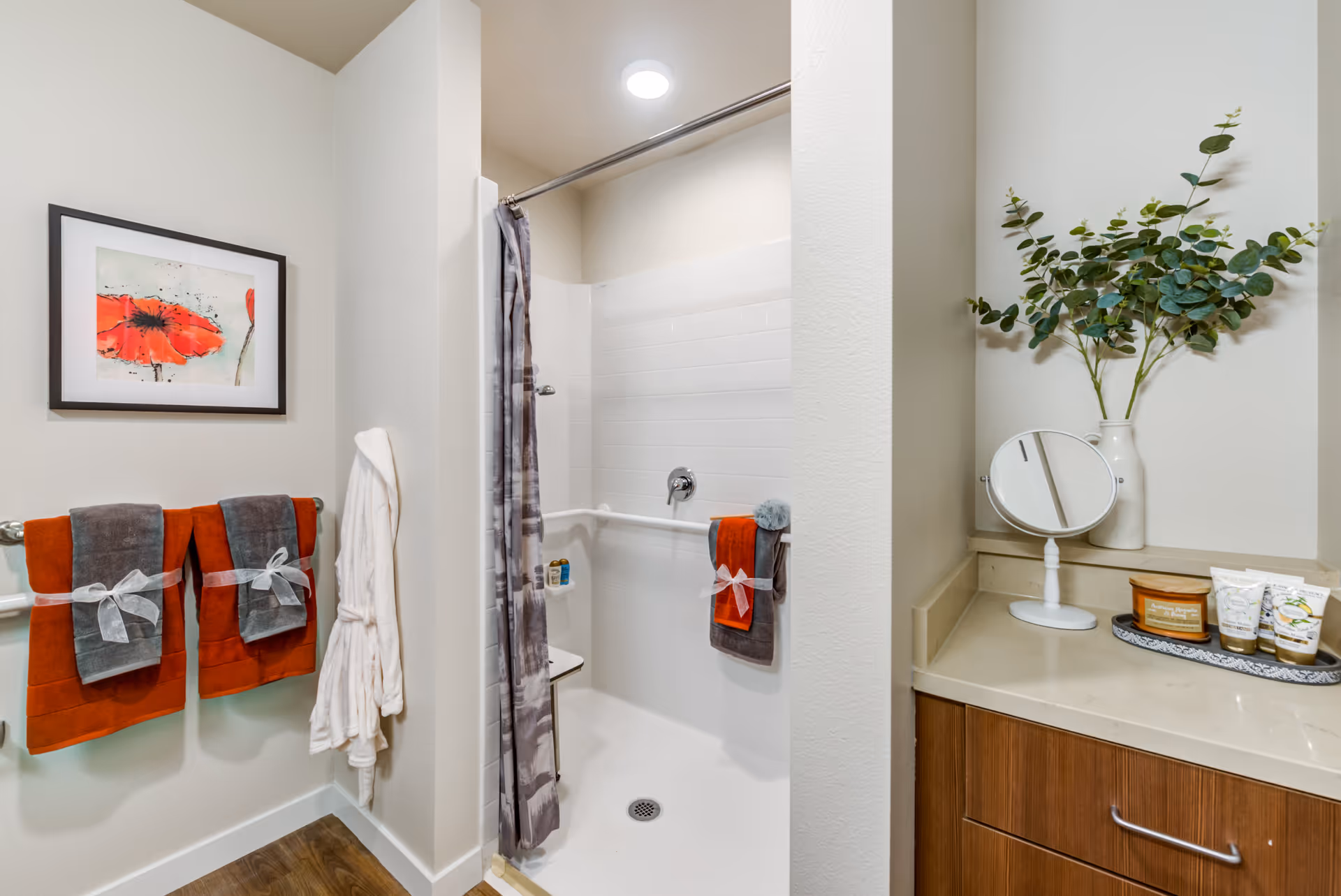 A bathroom with a walk-in shower featuring a gray and white shower curtain. Two sets of orange and gray towels are neatly hung on towel racks, each tied with a white ribbon. A white bathrobe hangs on a hook next to the towels. On the right side, there is a countertop with a small round mirror, a vase with green leafy branches, and a tray holding various toiletries. A framed picture of a red flower is mounted on the wall above the towels.