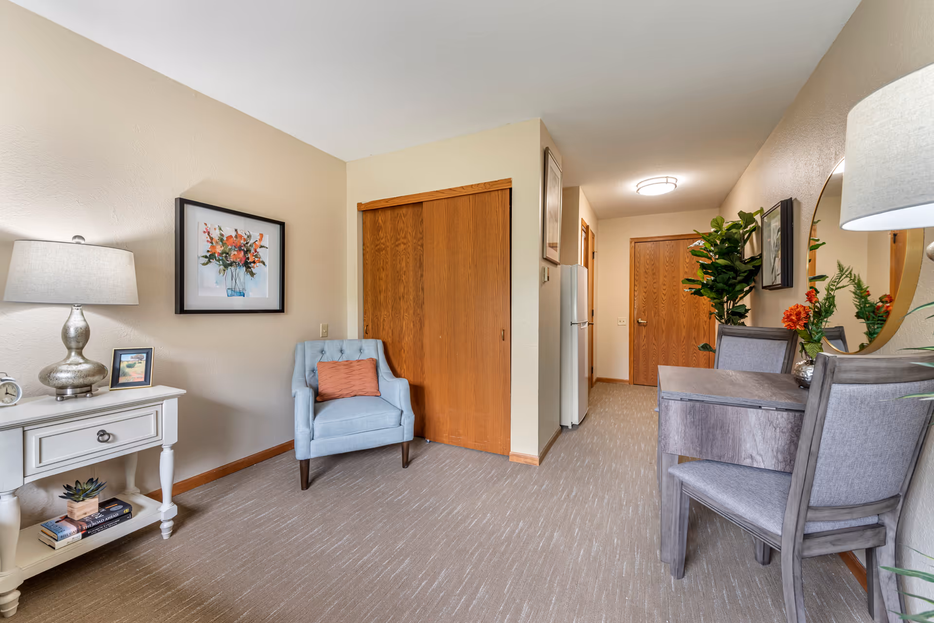 Interior view of a senior living facility room at Brookdale Faribault featuring a light blue armchair with an orange pillow, a white side table with a lamp and books, a wooden closet with sliding doors, a small dining table with two chairs, a round mirror on the wall, and decorative plants and artwork.