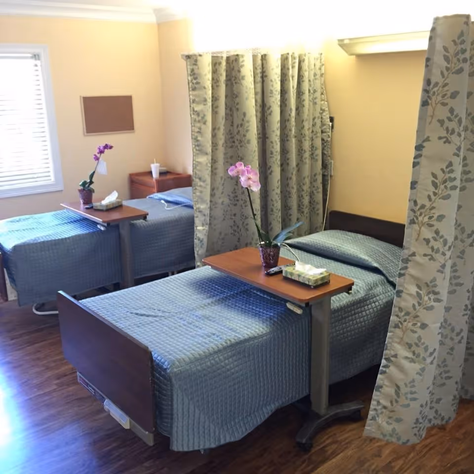 A room with two hospital-style beds separated by a floral privacy curtain. Each bed has a blue quilted bedspread and an overbed table with a potted orchid and a box of tissues. The room has wooden flooring, a window with blinds, and light yellow walls.