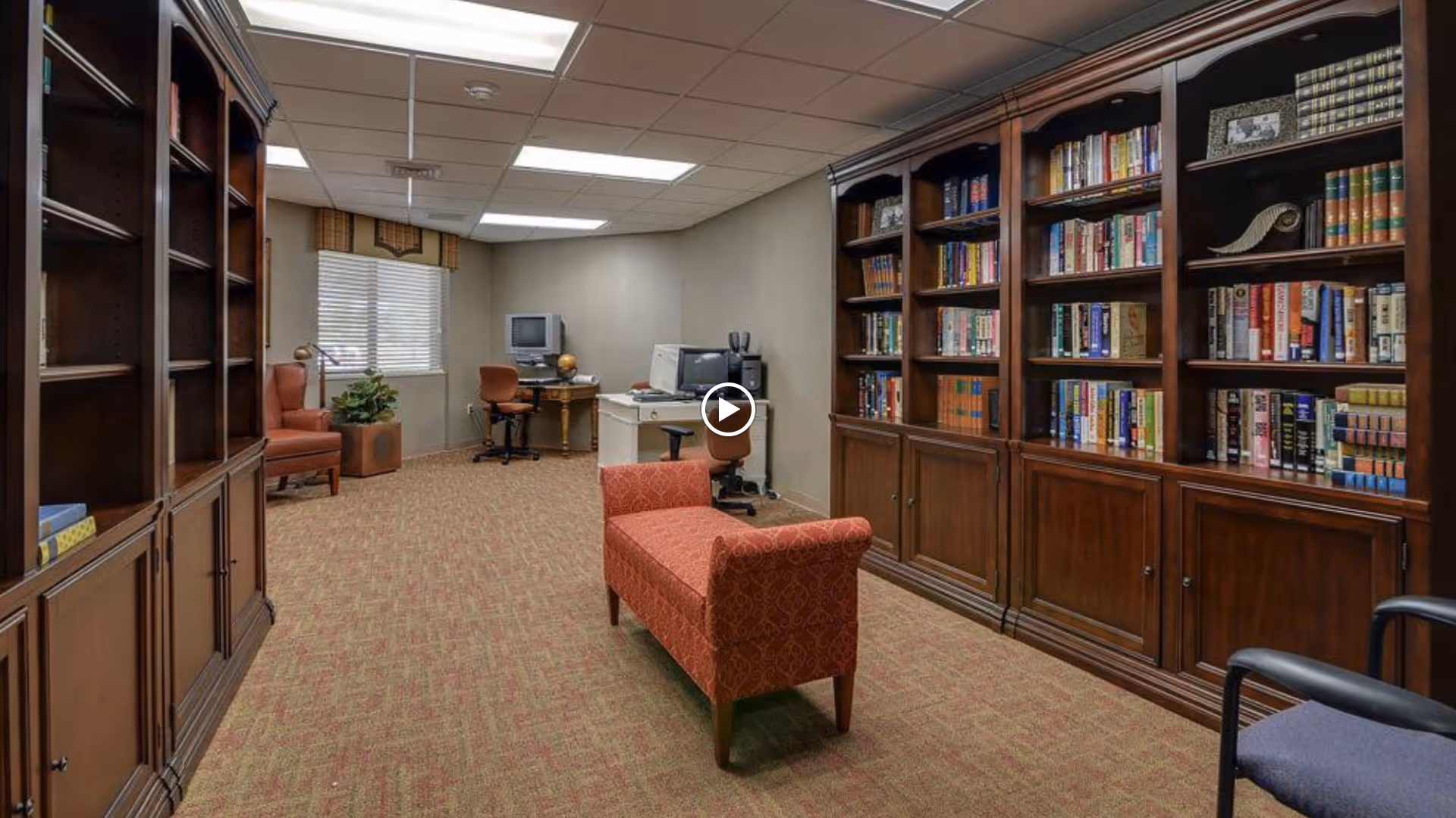 Carpeted community library or reading room with wooden bookshelves, seating, and a computer desk.